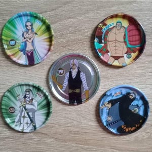 Può includere: Cinque gettoni da collezione circolari con personaggi anime. Ogni gettone presenta un'illustrazione di personaggio diversa con colori vivaci. I gettoni sono disposti su una superficie di legno marrone chiaro.