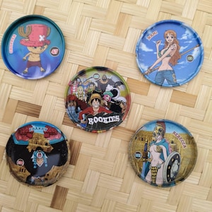 May include: Five colorful, circular collectible tokens featuring characters from the anime series One Piece. Each token displays a different character illustration, including Chopper, Nami, Franky, Rebecca, and a group of "Rookies".