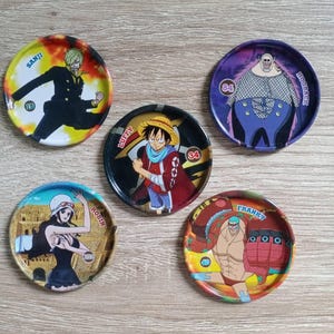 May include: Five circular collectible tokens with illustrations of characters from the anime One Piece. Each token features a different character, their name, and a number. The tokens are brightly coloured and set against a light wood grain background.