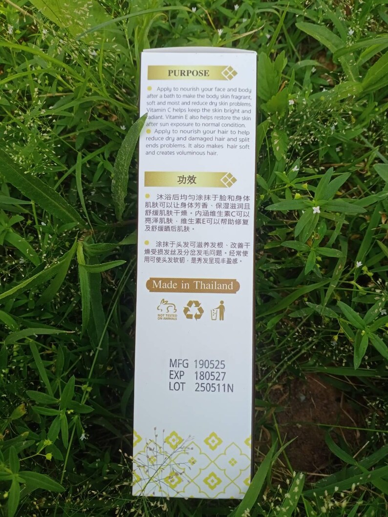 May include: A white product box with text in English and Chinese, set against a backdrop of green foliage. The box details the product's purpose, including nourishing skin and hair. The text "Made in Thailand" is also visible.
