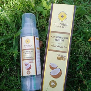May include: A clear spray bottle and a rectangular box of Pinnara Coconut Oil Serum, with the text "Coconut Oil Serum" and "Vitamin C&E". The bottle and box are on a bed of green grass. The bottle contains 85 ml.