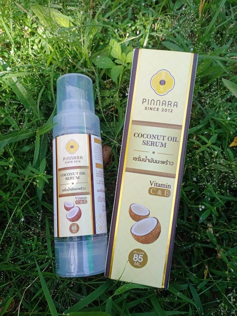 May include: A clear spray bottle and a rectangular box of Pinnara Coconut Oil Serum, with the text "Coconut Oil Serum" and "Vitamin C&E". The bottle and box are on a bed of green grass. The bottle contains 85 ml.