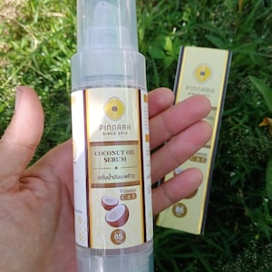 May include: A clear, cylindrical bottle of Pinnara Coconut Oil Serum, 85 ml, with a pump dispenser. The label features a gold and white design with text and a coconut illustration. The bottle is held against a backdrop of green grass.