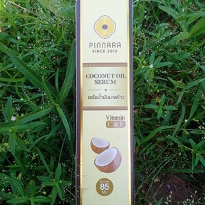 May include: A rectangular box of Pinnara Coconut Oil Serum, with text in English and Thai. The box is cream and brown, with a logo and the words "COCONUT OIL SERUM". It also features images of coconuts and states the serum contains Vitamin C & E. The box contains 85 mL.