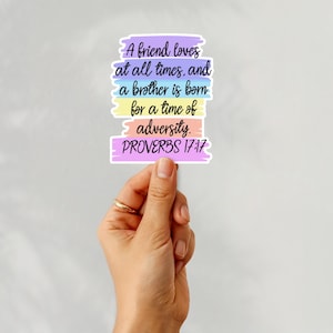 May include: A colorful sticker with the text "A friend loves at all times, and a brother is born for a time of adversity. PROVERBS 17:17". The sticker has a pastel color scheme and is held by a hand.
