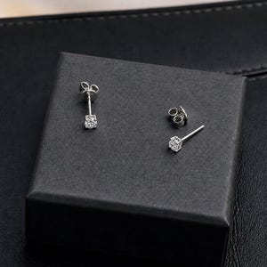 May include: A pair of silver stud earrings with a square, clear gemstone. One earring is displayed with its backing, and the other is shown separately. The earrings are on a black box, set against a dark background.