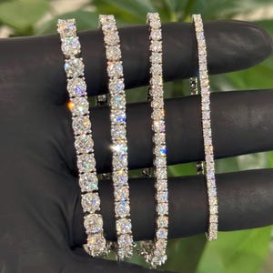 May include: Four silver-toned tennis bracelets featuring round-cut clear stones arranged in a continuous line. The bracelets vary in width, with the leftmost being the thickest and the rightmost being the thinnest, all displaying high-shine metallic settings.