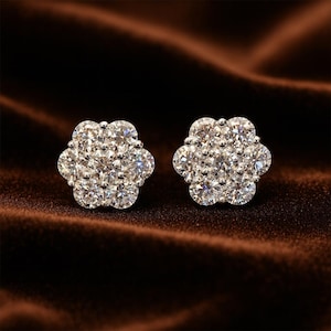 May include: A pair of flower-shaped stud earrings. Each earring is composed of multiple small, round, clear gemstones clustered together to form a floral design. The earrings are set against a dark brown, velvety background.
