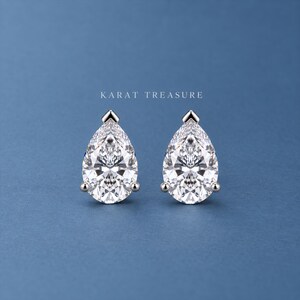May include: A pair of pear-shaped stud earrings featuring clear, faceted stones set in silver-toned metal prongs. The text KARAT TREASURE is centered above the earrings. Each earring has a pointed top and a rounded base, reflecting light.