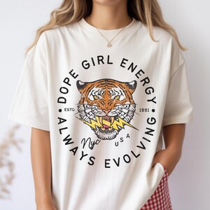 Puede incluir: Camiseta color crema con un gráfico de tigre y el texto "DOPE GIRL ENERGY ALWAYS EVOLVING NYC USA ESTD 1981". El tigre tiene rayas naranjas y negras con un rayo amarillo. La persona lleva un pantalón corto a cuadros rojos y blancos.