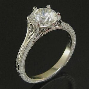 May include: A silver engagement ring with a large, clear, round-cut diamond. The ring features intricate filigree detailing along the band and around the diamond setting. The ring is set against a dark background.