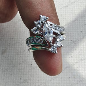 May include: A silver-toned ring set featuring a central marquise-cut clear stone surrounded by multiple smaller round-cut clear stones. The band has a curved, layered design with additional round-cut stones embedded along the metal surface.