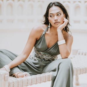 Könnte beinhalten: Ein salbeigrüner Jumpsuit mit Neckholder-Ausschnitt und weiten Beinen. Die Taille ist mit einem Perlendesign verziert. Das Model trägt weiße Armbänder und Ohrringe. Der Jumpsuit ist aus fließendem Stoff gefertigt.