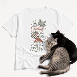 May include: A white t-shirt featuring a graphic design with the text I'm Falling For FALL, a leopard print leaf, a checkered pumpkin, and a mushroom. A grey tabby cat and a black cat are resting on the lower right side of the shirt fabric.