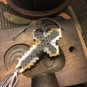May include: A crocheted cross ornament featuring a color palette of dark gray, light gray, white, and tan. The cross has a scalloped edge and includes a braided gray hanging loop at the top and a multi-colored tassel at the bottom point.