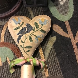 May include: A heart-shaped, beige fabric ornament with a cross-stitched design of a blue bird perched on a flowering branch. The ornament is attached to a spool of thread with a green and pink ribbon. The background is a dark, patterned fabric.