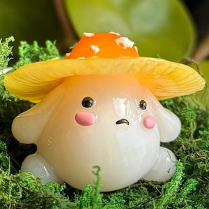 May include: A whimsical, mushroom-shaped figurine with a yellow and orange cap, white spots, and a sad expression. The figurine has a round body, small arms, and pink cheeks. It is set in a bed of green moss.