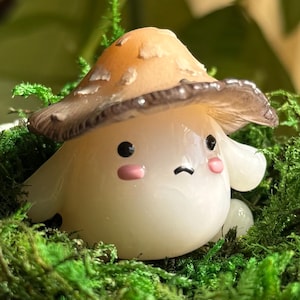 May include: A whimsical mushroom figurine with a light brown and tan cap, black eyes, and pink cheeks, nestled in vibrant green moss. The mushroom has a cheerful, cartoonish appearance. The background is blurred, suggesting a natural setting.