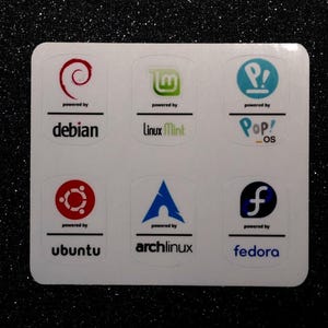 May include: A white sticker sheet with logos for six operating systems: Debian, Linux Mint, Pop!_OS, Ubuntu, Arch Linux, and Fedora. Each logo has the text "powered by" beneath it.