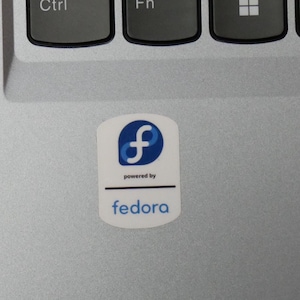 May include: A white sticker featuring a blue and white "f" logo, with the text "powered by" and "fedora" in a sans-serif font. The sticker is affixed to a silver laptop surface, near the keyboard.