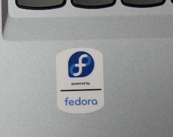 Può includere: Un adesivo bianco con un logo "f" stilizzato blu e bianco, con le parole "powered by" e "fedora" in un font sans-serif. L'adesivo è su una superficie argentata di un laptop, vicino alla tastiera.