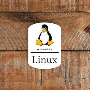 Pode incluir: Um adesivo branco com o logotipo do Linux, um pinguim de desenho animado com corpo preto, barriga branca e pés e bico amarelos. O texto "powered by" está acima de uma linha horizontal, com "Linux" abaixo em negrito, sem serifa. O fundo é uma superfície de madeira.
