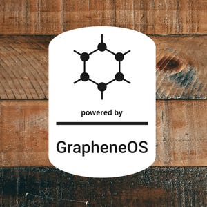 May include: A white oval logo with a black molecular structure graphic and the text "powered by" above a horizontal line. Below the line is the text "GrapheneOS" in a bold, sans-serif font. The background is a wooden surface.