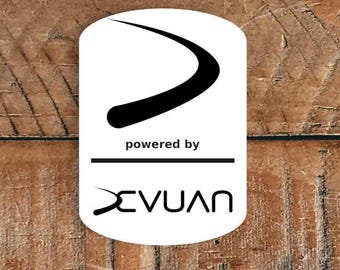 May include: White, rectangular sticker featuring a black curved design and the words "powered by" above the name "XEVUAN" in black. The sticker is on a wooden surface.