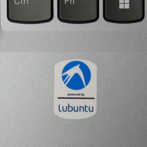 May include: A white sticker with a blue logo and the text "powered by Lubuntu" is affixed to a silver laptop. The logo features a stylized bird within a blue circle. The sticker is rectangular with rounded corners.