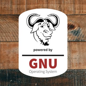 May include: A white rectangular sticker with rounded corners featuring a black line drawing of a gnu head at the top. Below the gnu, the text reads "powered by" in black, followed by a horizontal line and the red text "GNU" and "Operating System".