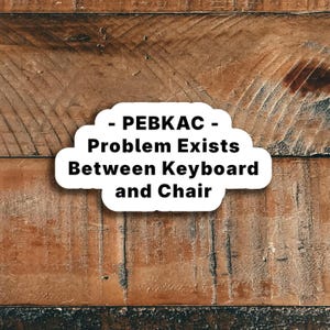 May include: A white sticker with the text "- PEBKAC - Problem Exists Between Keyboard and Chair" in black, set against a textured, brown wooden background. The sticker has a cloud-like shape.