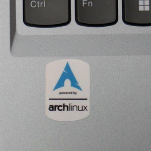Peut inclure: Un autocollant blanc avec un logo bleu et le texte "powered by archlinux" est collé sur un ordinateur portable argenté. Le logo est une forme bleue abstraite au-dessus du texte. L'autocollant est de forme ovale.