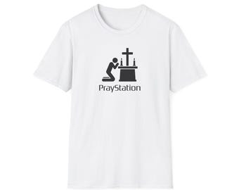 May include: A white short-sleeved t-shirt featuring a black graphic of a person kneeling in prayer before an altar with a cross and two candles. Below the graphic, the text PrayStation is printed in a black sans-serif font.