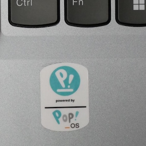 May include: A white sticker with the text "Pop!_OS" in teal and the words "powered by" above it. The sticker is on a silver laptop surface, near the Ctrl and Fn keys.