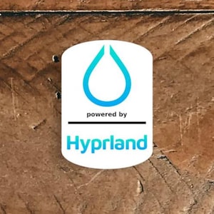 Puede incluir: Un logotipo ovalado blanco con un diseño de lágrima azul y el texto "powered by" encima de una línea negra. Debajo de la línea está la palabra "Hyprland" en azul y verde.