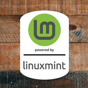 May include: A white sticker with a green circle containing a stylized 'lm' logo in gray. Below the logo, the text reads 'powered by' and 'linuxmint' in a sans-serif font. The background is a wooden surface.