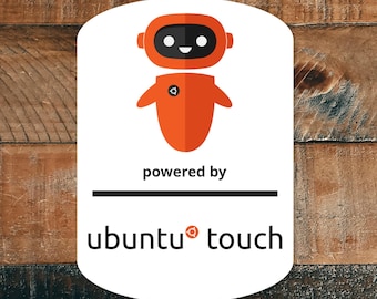 Può includere: Un adesivo bianco con un robot arancione e la scritta "powered by ubuntu touch". Il robot ha una faccia nera e un piccolo disegno circolare sul petto. L'adesivo è su uno sfondo di legno.