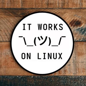 Può includere: Un adesivo circolare bianco con testo nero che recita "IT WORKS" sopra un'emoticon e "ON LINUX". L'adesivo ha un bordo nero ed è posto su uno sfondo di legno.