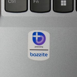 May include: A white sticker with a blue and purple logo and the text "powered by" above the word "bazzite" in a sans-serif font. The sticker is on a silver laptop surface, near the keyboard.