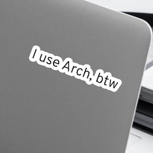 Peut inclure: Autocollant blanc avec le texte "I use Arch, btw" sur un ordinateur portable gris. L'autocollant est légèrement incliné et l'ordinateur portable est partiellement visible. Le texte est en police sans-serif.
