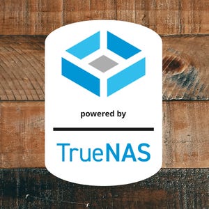 Puede incluir: Un logotipo ovalado blanco con un diseño geométrico azul y el texto "powered by" encima de una línea negra. Debajo de la línea está el texto azul "TrueNAS". El fondo es una superficie de madera.