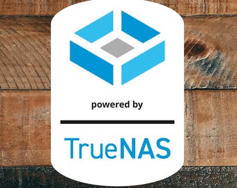 Può includere: Un logo ovale bianco con un design geometrico blu e la scritta "powered by" sopra una linea nera. Sotto la linea c'è il testo blu "TrueNAS". Lo sfondo è una superficie di legno.