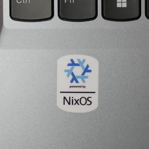 May include: A white sticker with the text "powered by" above "NixOS" and a blue and light blue geometric design. The sticker is on a silver laptop surface.