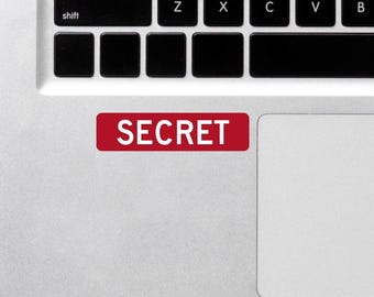 Può includere: Un adesivo rettangolare rosso con la scritta "SECRET" in lettere bianche è posizionato su un laptop argentato. L'adesivo è posizionato vicino alla tastiera e al touchpad.