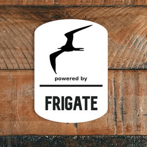 Może przedstawiać: Biały, zaokrąglony prostokąt z czarną sylwetką ptaka w locie. Pod ptakiem znajduje się tekst "powered by" nad słowem "FRIGATE" pogrubioną, czarną czcionką. Tło to teksturowana, brązowa powierzchnia drewniana.