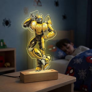 May include: A yellow and black Bumblebee robot night light with the name "NOAH" on the chest. The light is on a wooden base and is plugged in. The background shows a child sleeping in a bed with a Spider-Man comforter.