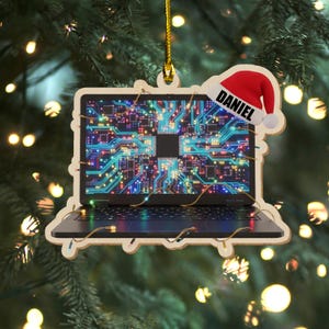 K&ouml;nnte beinhalten: Weihnachtsbaumschmuck in Form eines Holz-Laptops mit einem Leiterplatten-Design auf dem Bildschirm. Der Schmuck hat einen Weihnachtsmannhut mit dem Namen "DANIEL". Der Schmuck h&auml;ngt an einem Weihnachtsbaum mit Lichtern.