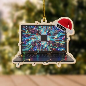K&ouml;nnte beinhalten: Weihnachtsbaumschmuck aus Holz in Form eines Laptops mit einem Leiterplattendesign auf dem Bildschirm. Der Schmuck hat einen Weihnachtsmannhut mit dem Namen "DANIEL". Der Laptop ist mit kleinen Lichtern verziert.