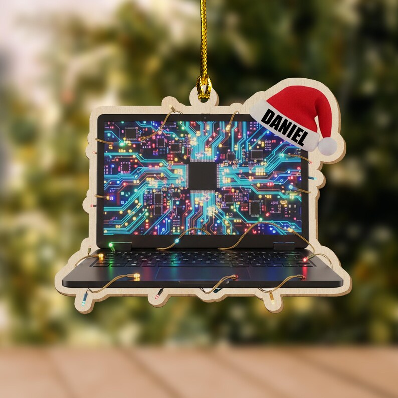 K&ouml;nnte beinhalten: Weihnachtsbaumschmuck aus Holz in Form eines Laptops mit einem Leiterplattendesign auf dem Bildschirm. Der Schmuck hat einen Weihnachtsmannhut mit dem Namen "DANIEL". Der Laptop ist mit kleinen Lichtern verziert.