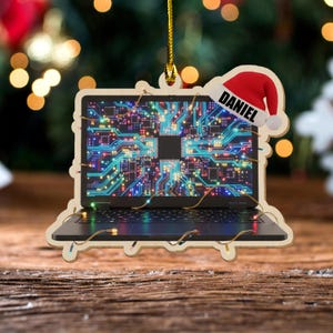 K&ouml;nnte beinhalten: Weihnachtsornament in Form eines Holz-Laptops mit einem Leiterplatten-Design auf dem Bildschirm. Das Ornament hat einen Weihnachtsmannhut mit dem Namen "DANIEL". Das Ornament ist mit kleinen Lichtern verziert.